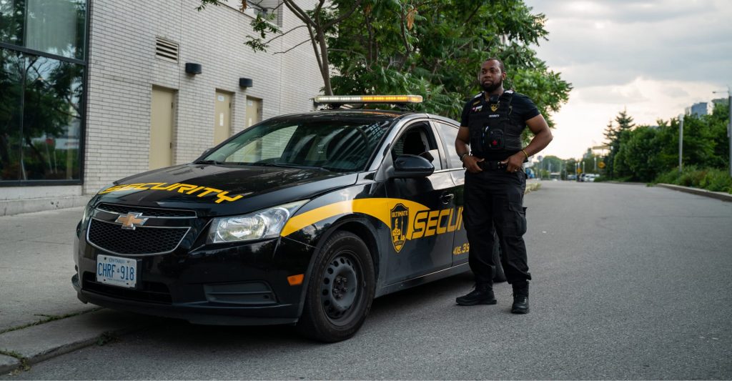 A security officer from the Special Response Unit stands next to a black and yellow security patrol car on a quiet street.
