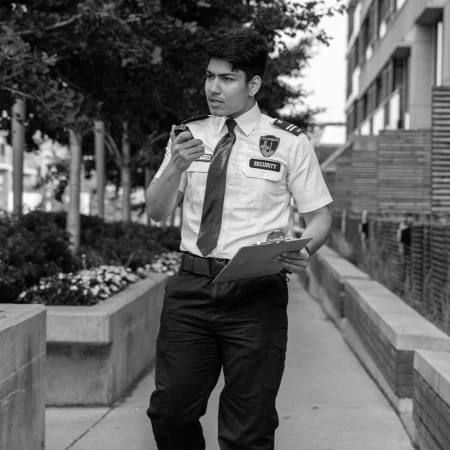 A security guard in uniform patrols an outdoor path, clipboard in hand and walkie-talkie at the ready, showcasing the professional security guard services in Toronto amidst a backdrop of buildings and trees.