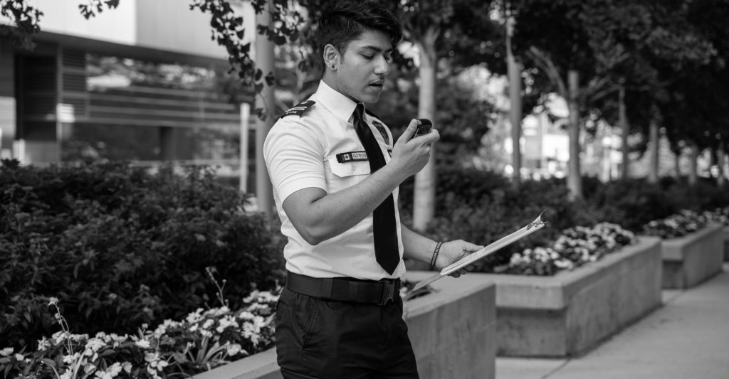 A private security man in uniform standing on a sidewalk with a cell phone.