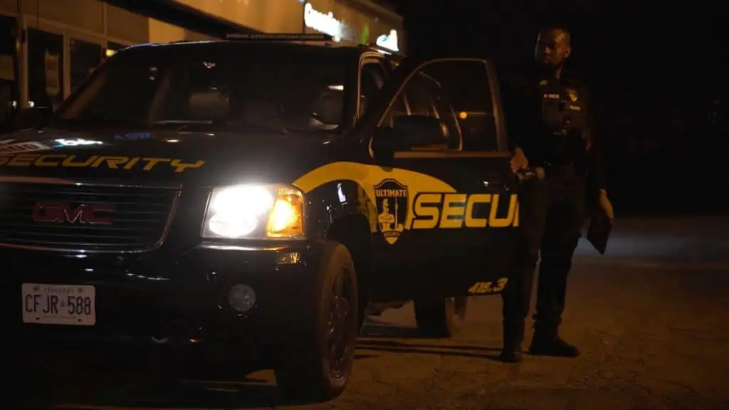 A mobile security guard is standing in front of a GMC truck at night.