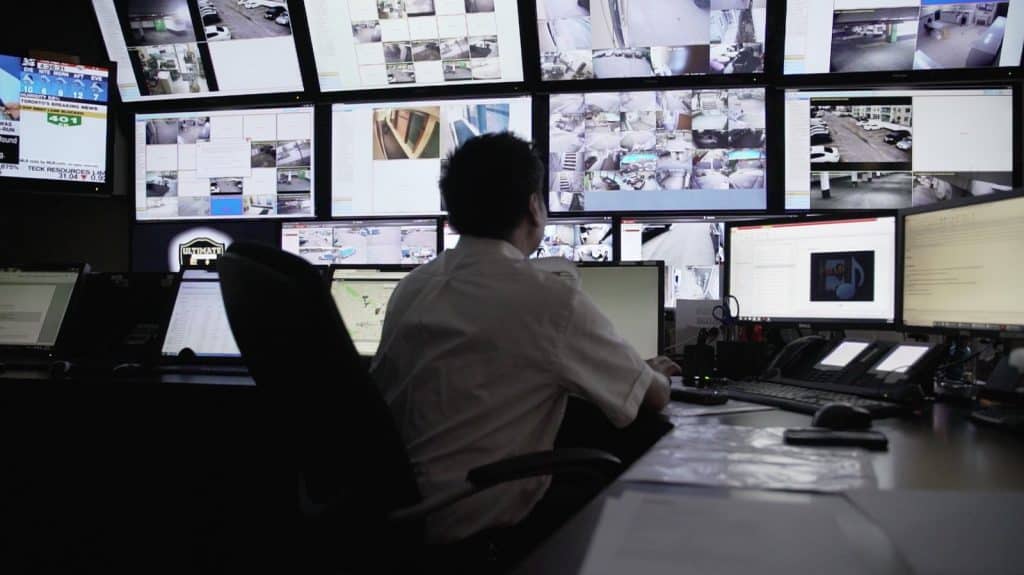 A man is sitting at a computer screen in an apartment with multiple monitors, reviewing home security footage.
