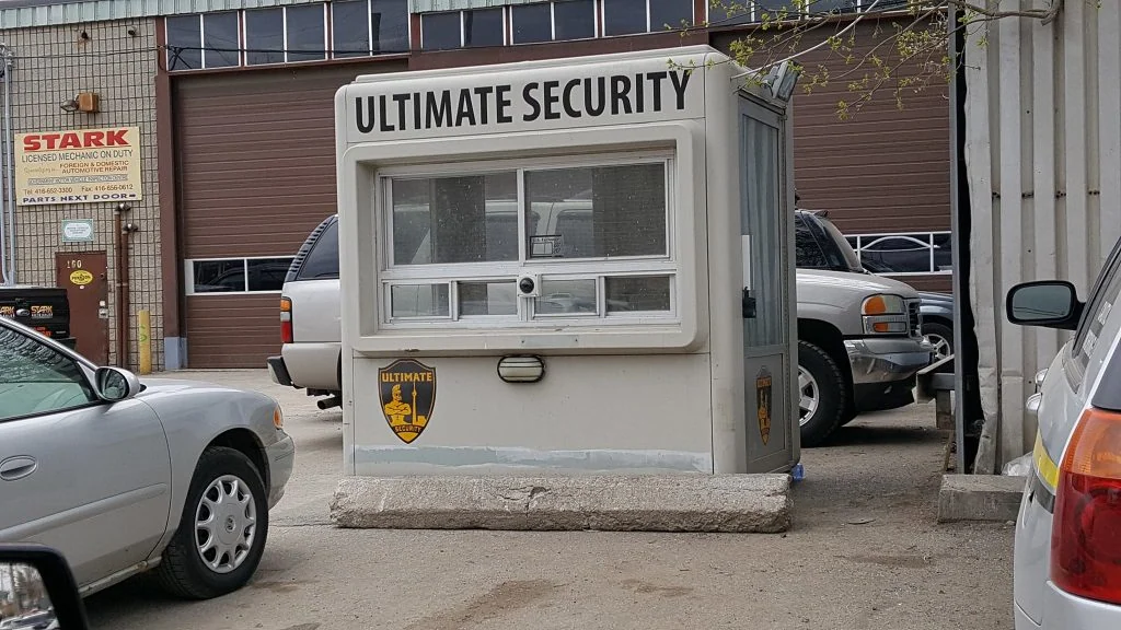 A security booth labeled "ultimate security" featuring an access control system in a parking lot, with parked cars and an industrial building in the background.