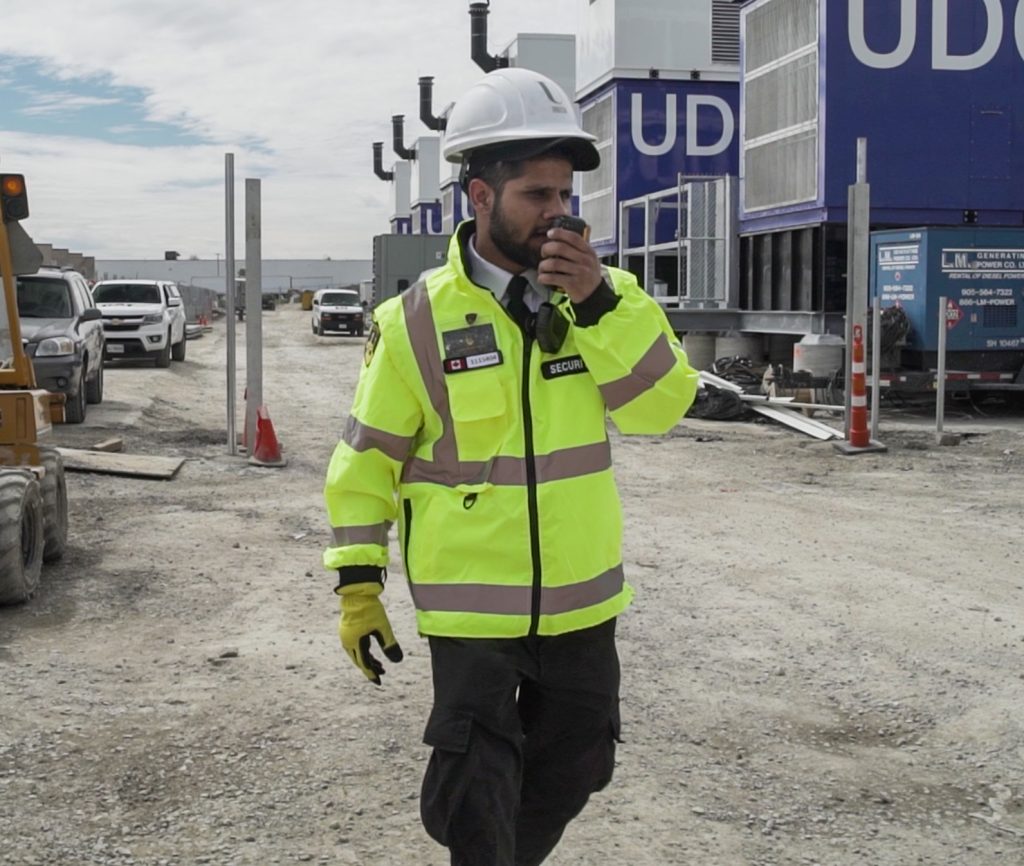 323634672 2369256193242532 3614081929298318388 n A Neighborhood Watch member in a high-visibility jacket and helmet, talking on a walkie-talkie at a construction site.