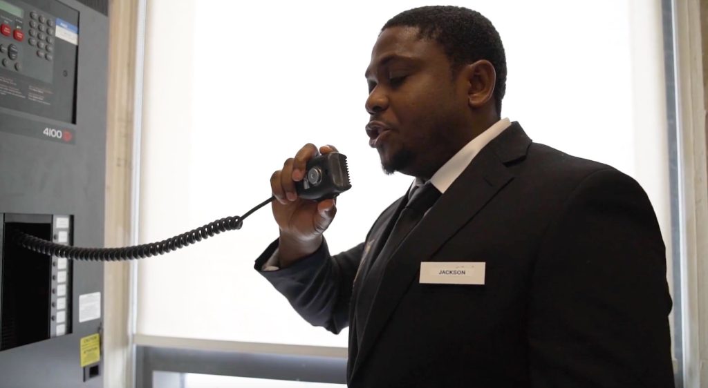 A male security guard named Jackson speaking into a walkie-talkie in an elevator, coordinating alarm monitoring.