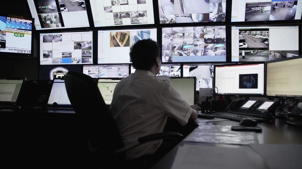 A person in a white shirt monitors multiple screens displaying various surveillance views in a dimly lit access control room.