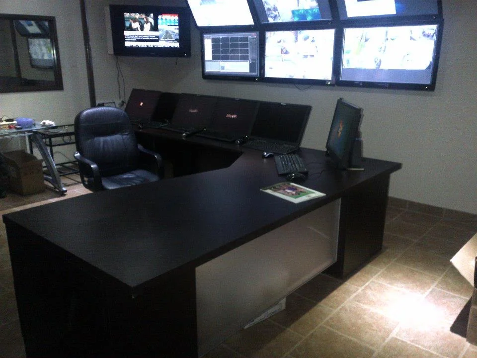 A modern office featuring a large desk with multiple laptops and a wall-mounted array of television screens showing various channels, including dedicated screens for alarm monitoring.