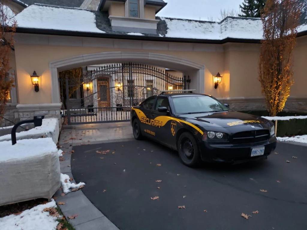 A black and yellow Residential Security Services patrol car parked in front of a gated entrance to a large residential home with snow on the ground.