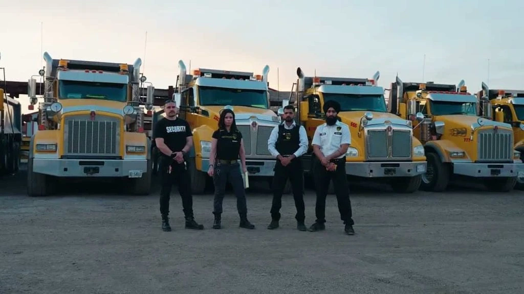 Four truck drivers stand confidently in front of a row of parked semi-trucks during dusk, accompanied by security guard services.