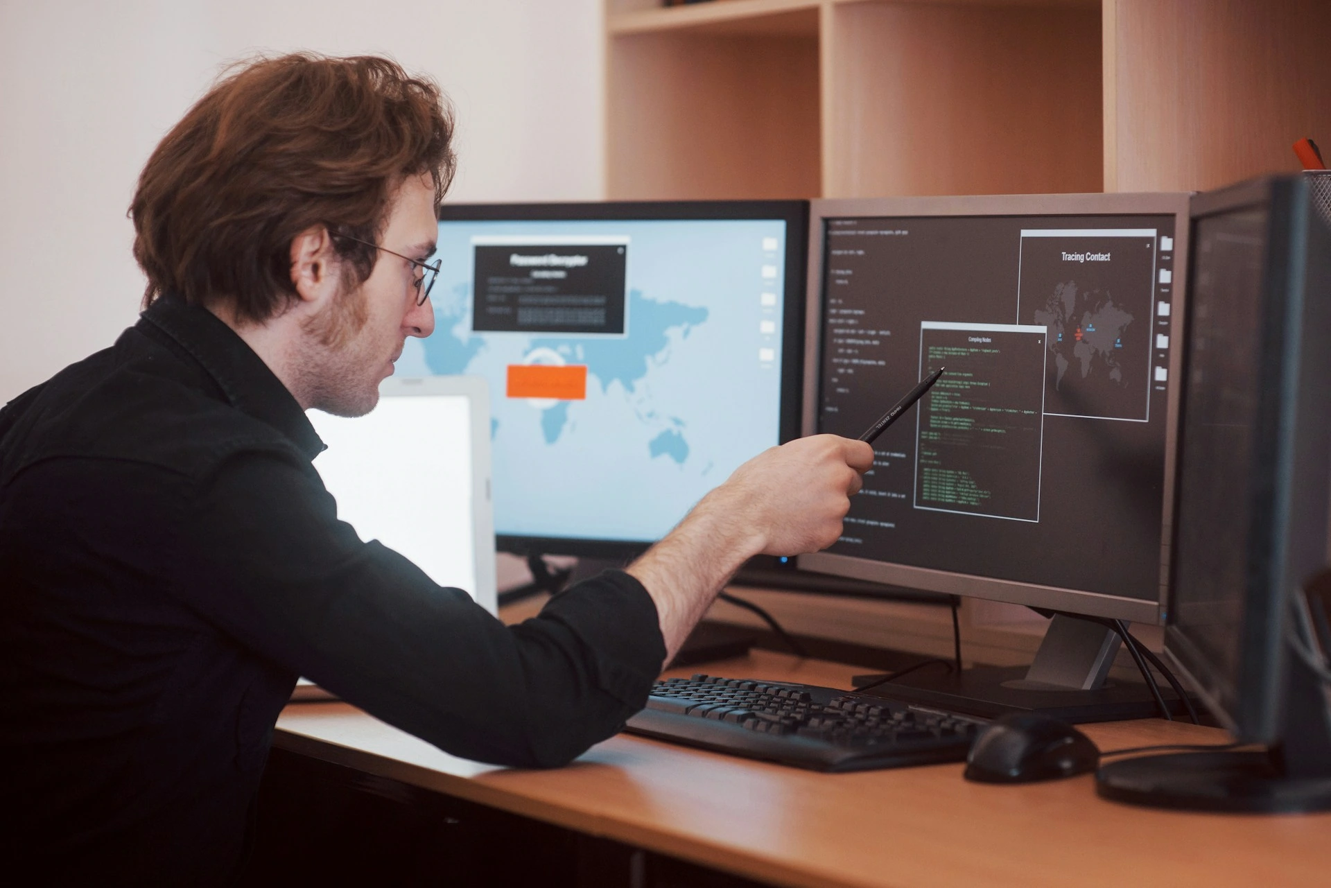 A person in a black shirt points at a computer screen displaying code, illustrating key cybersecurity tips. Two other monitors sit on the desk, one showing a world map, hinting at global data protection strategies.