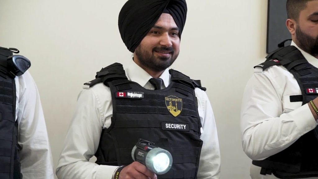 A Sikh security guard in a bulletproof vest holds a flashlight and smiles, standing beside a colleague in a similar uniform.