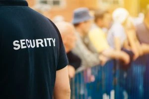 Event security staff member in a black shirt labeled "security" overseeing a crowd behind metal barriers, with a focus on the uniform's text.