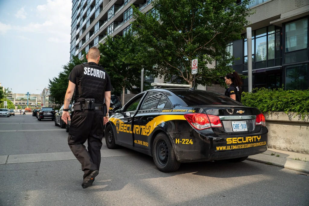 Two security officers walk near a parked security car on a city street next to a modern residential building, ensuring the area aligns with the local crisis management plan.