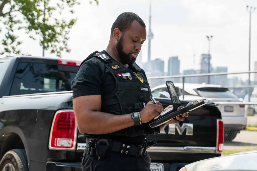 A security officer in uniform writes on a notepad near a black truck in a parking lot, as the city skyline looms in the background. Ever vigilant, these security consultants ensure every detail aligns with urban safety protocols.