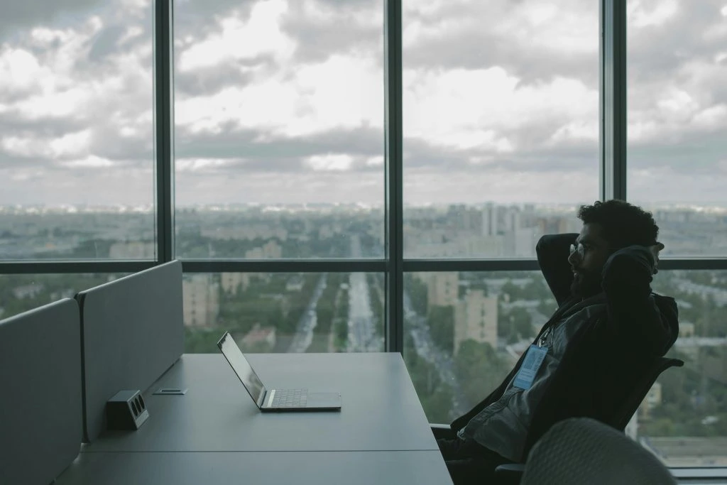 A person sits at a desk with a laptop in front of them, looking out a large window with a cityscape view. They seem deep in thought, perhaps pondering cybersecurity solutions for high-profile clients, with their hands resting behind their head.