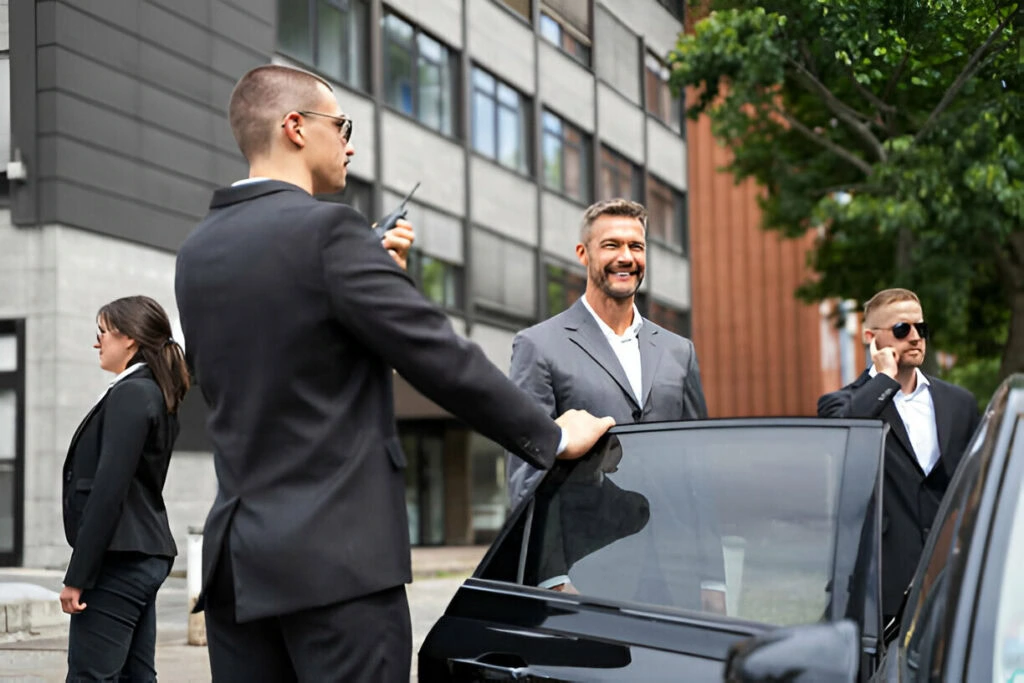 Four individuals in suits stand near a car in a modern urban setting. Three have earpieces and one holds a two-way radio, clearly indicating executive protection services. The fourth person appears to be their focus.