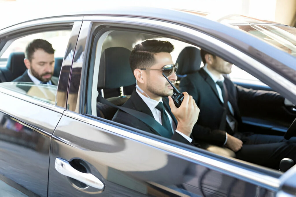 Three men in suits sit inside a car; the driver is on a radio, the front passenger looks ahead attentively, and the rear passenger is checking his phone. The scene exudes professionalism, highlighting the precision of executive protection services.