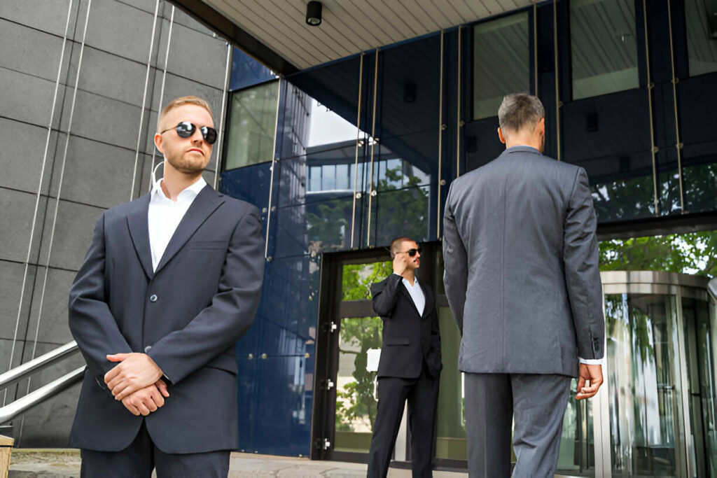 Three men in suits and sunglasses stand outside a modern building with glass walls. One faces forward, one is on the phone, and one faces away, exemplifying the precision of Executive Protection.