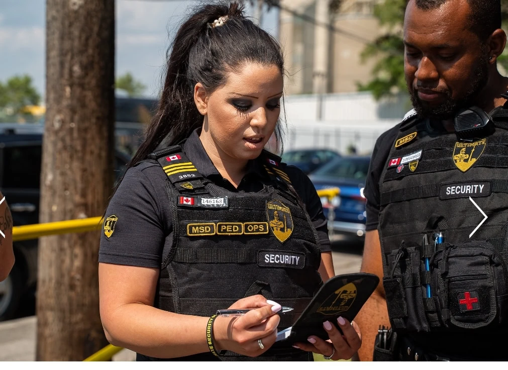 Two security guards in uniform are discussing information on a tablet. Both wear black vests with various patches and are standing outdoors, near a wooden pole and yellow tape.