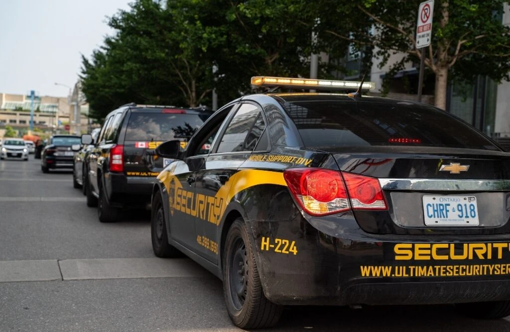 Two black security vehicles with yellow markings are parked by the roadside, likely belonging to security guards. The license plate of the nearest vehicle reads "CHRF-918." Buildings and trees are visible in the background.