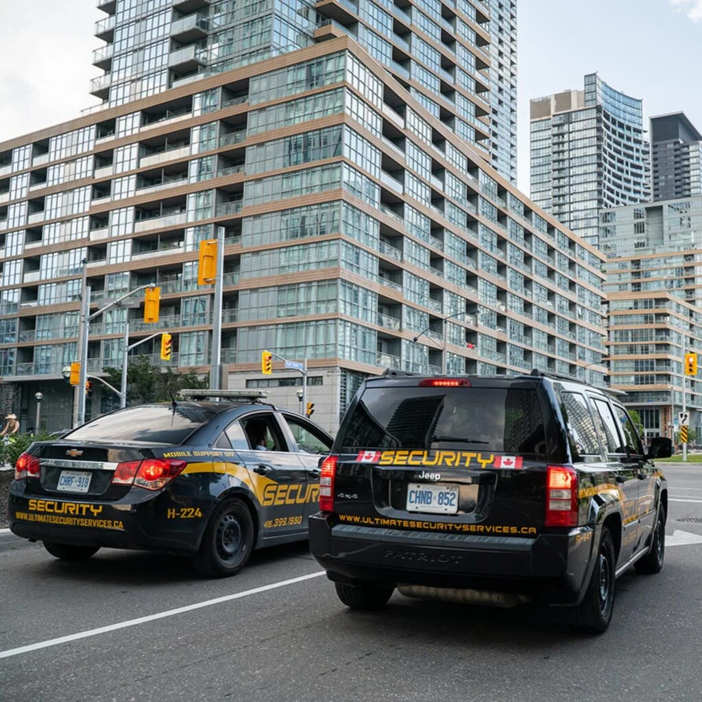 Two security vehicles are parked on a city street corner in front of modern high-rise buildings, serving as part of the neighbourhood watch.