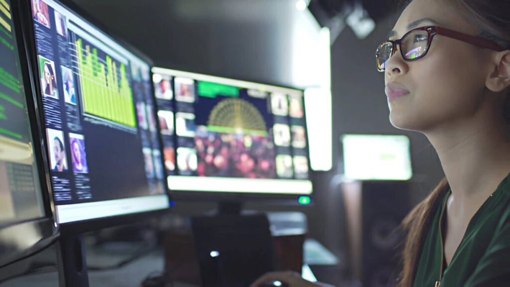 A person with glasses is using multiple computer screens displaying data charts and photos in a dimly lit room, meticulously analyzing data as part of Event Security Solutions for High-Profile Clients.