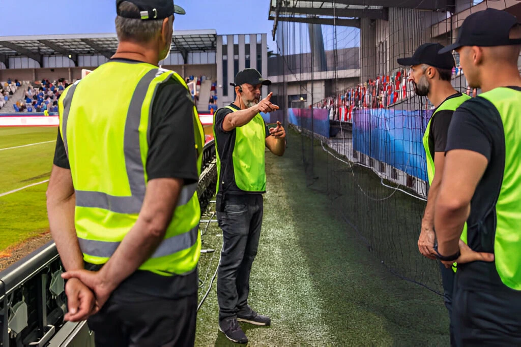 A man in a yellow vest gestures while speaking to three other men in yellow vests near a sports field, providing event security for high-profile clients, with scattered spectators visible in the background.