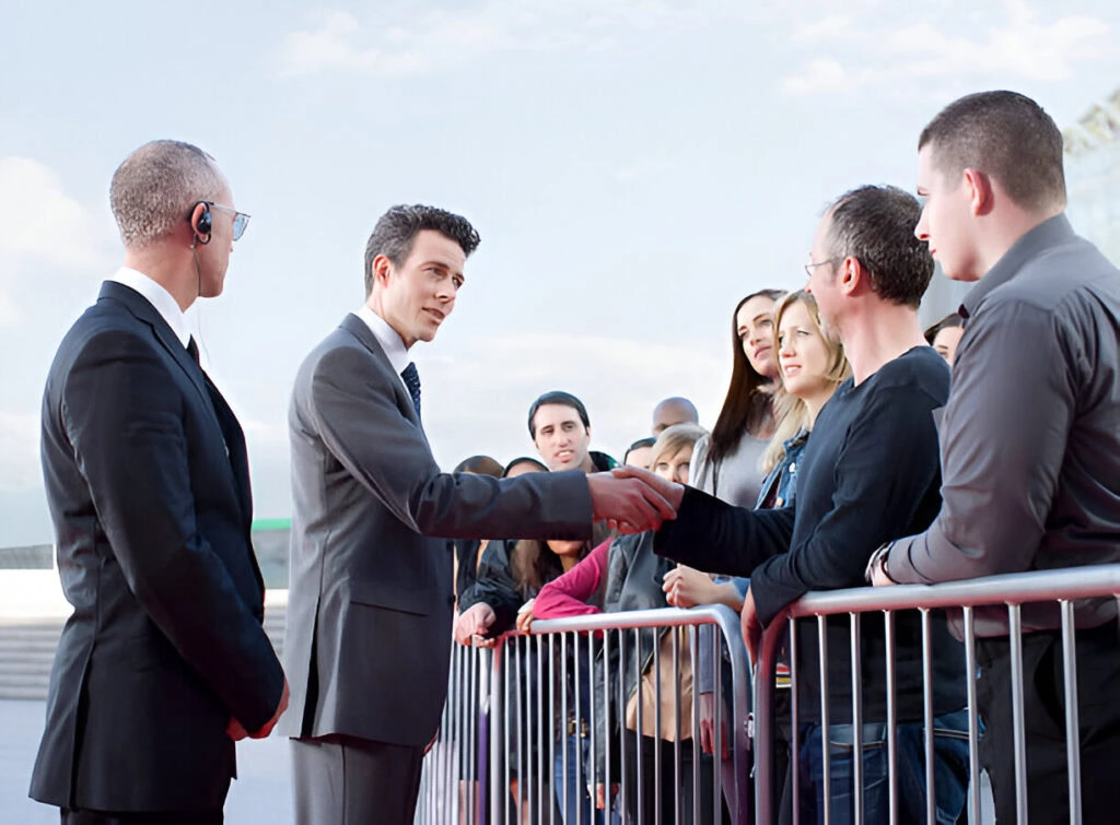 A man in a suit is shaking hands with a person in the crowd behind a barrier, while another man in a black suit with an earpiece from Event Security Solutions for High-Profile Clients stands nearby. Several people are watching.