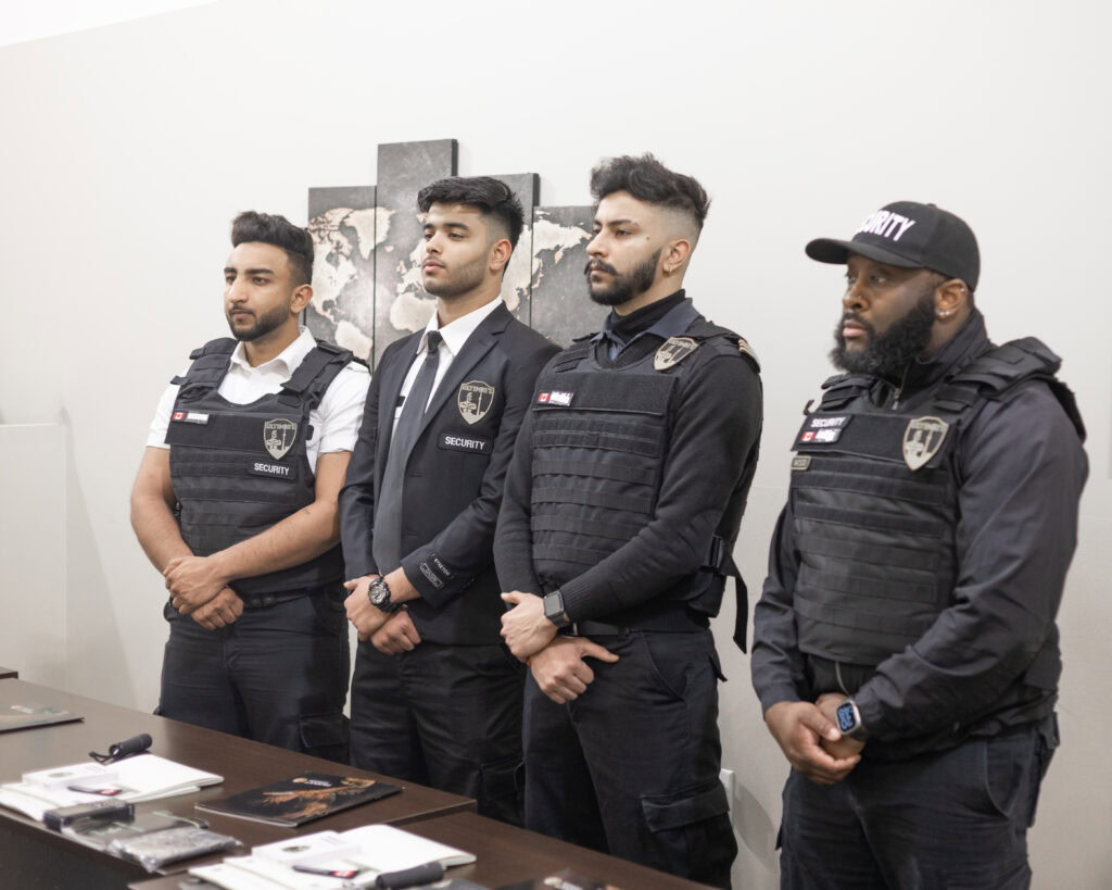 Four security guards wearing uniforms and badges stand in a line, facing forward. Three wear tactical vests; one wears a suit jacket. Various items are placed on a table in front of them, showcasing the professional security guard services in Toronto.