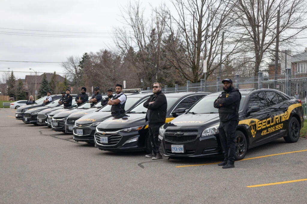 A group of security personnel stands with arms crossed in front of a row of black security vehicles in a parking lot. The cars, emblazoned with the Mobile Security Services logo and contact number, ensure peace and safety.