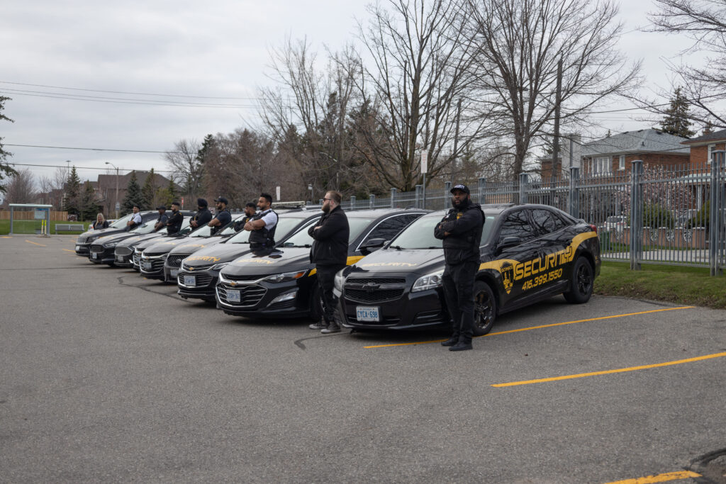 A line of dedicated security personnel from our mobile security services stands in front of parked vehicles labeled with "security" in a parking lot near residential houses.