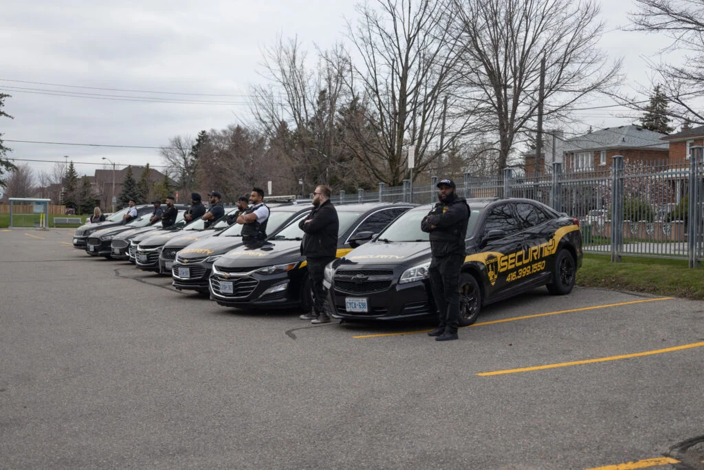 A line of dedicated security personnel from our mobile security services stands in front of parked vehicles labeled with "security" in a parking lot near residential houses.