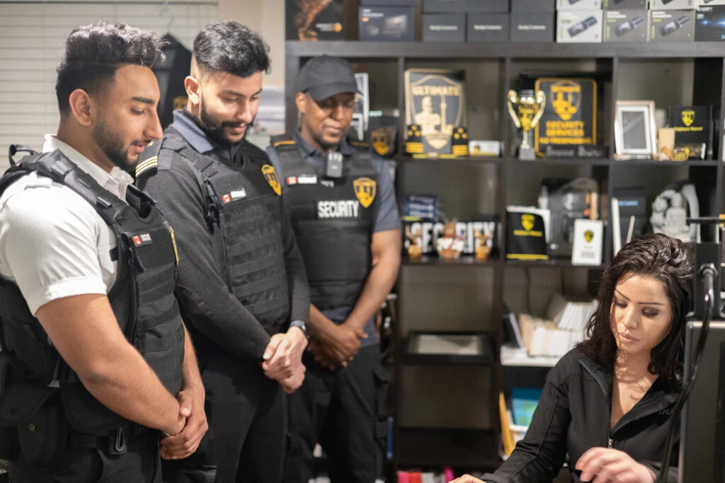 Four security officers are standing, wearing uniforms, as a woman seated at a desk works on paperwork in an office filled with awards and security equipment. This scene exemplifies the professionalism expected from top-tier security services in Toronto.