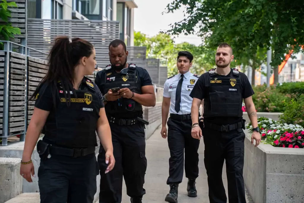 Four security guards stroll down a pathway, clad in black uniforms, with one distinctively standing out in white. Trees and buildings frame the background, adding a touch of nature to the scene.