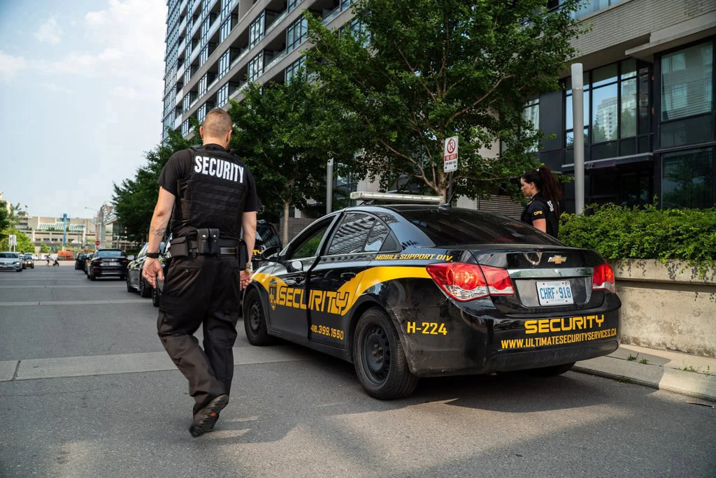 Two security guards stand vigilantly by a security car on a bustling city street, framed by a towering building and lush trees in the background.