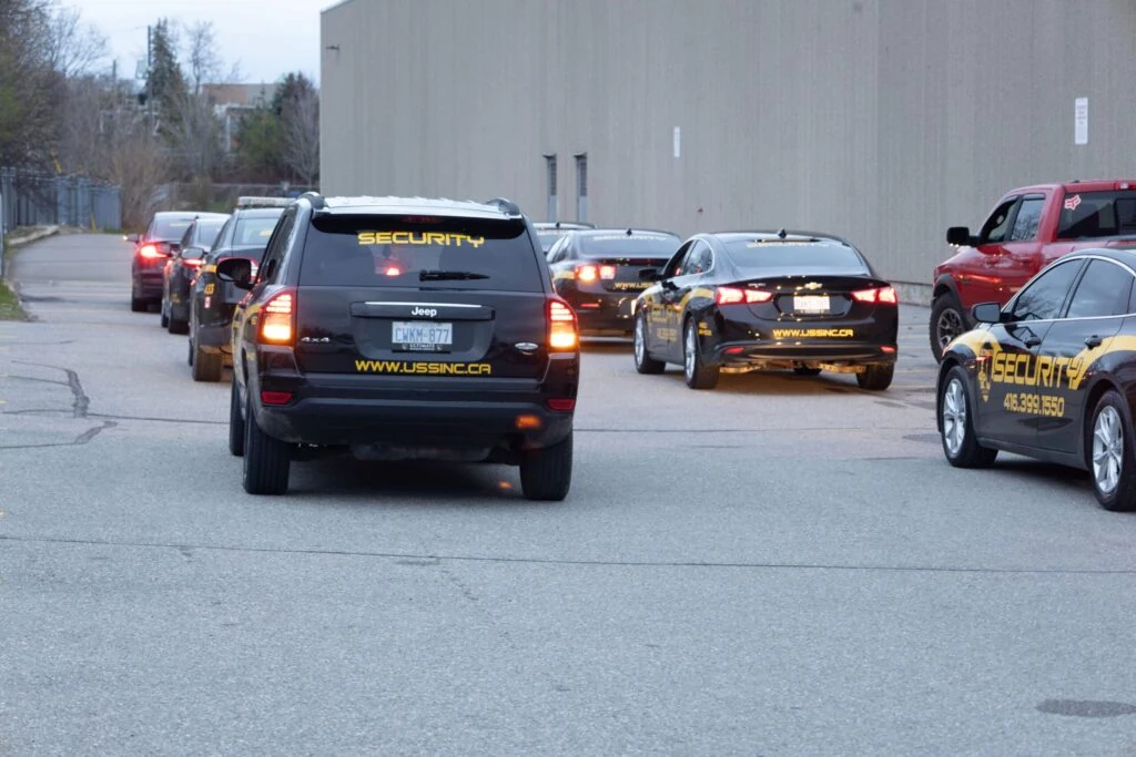A line of security vehicles with "SECURITY" markings, part of mobile security services, drives down a paved road next to a large beige building.