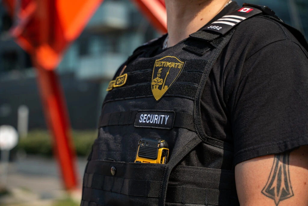 A security guard wearing a black vest labeled "Security" and holding a yellow device stands outdoors near a modern building, resembling a private detective in Toronto on surveillance duty.