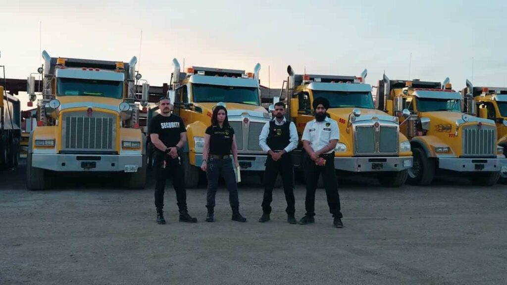 Four security personnel stand in front of five yellow dump trucks parked on a gravel lot at dusk, ensuring strong site security.