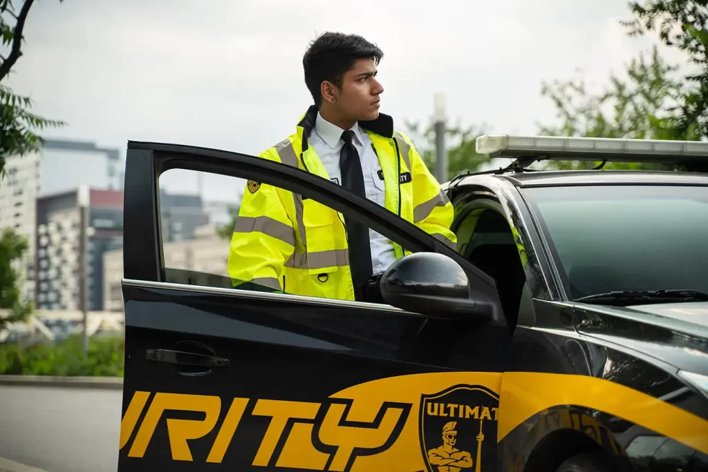 A security guard in a yellow jacket stands by the open door of a black and yellow mobile security services vehicle, looking to the side.