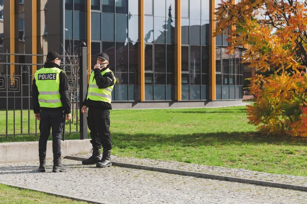 Two police officers in black uniforms and yellow reflective vests, part of the neighborhood watch, stand on a cobblestone path talking, with a modern glass building and a tree with autumn leaves in the background.