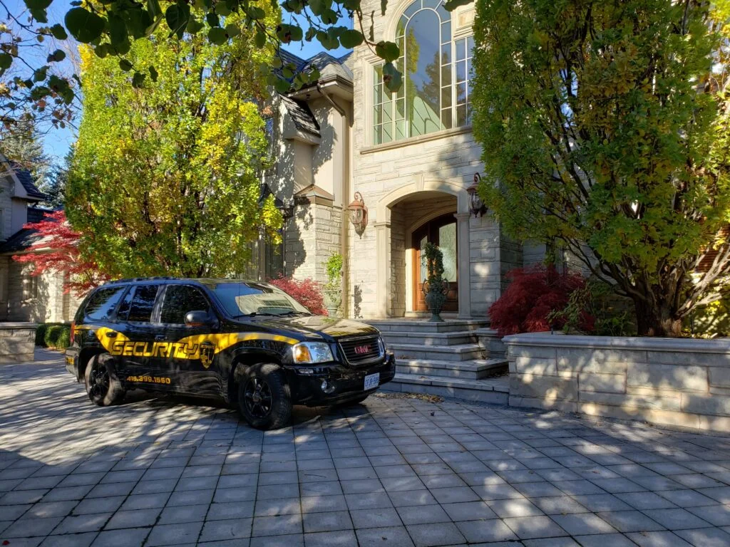 A black GMC SUV with "Security" markings, part of a neighborhood watch in Canada, is parked on a stone driveway in front of a large, two-story house surrounded by trees and shrubs.