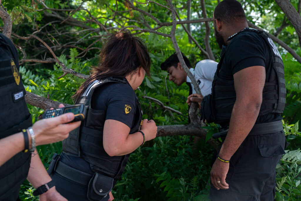 Three people in black uniforms inspect a man partially concealed by foliage, while another person holds a device nearby in an outdoor, wooded area.