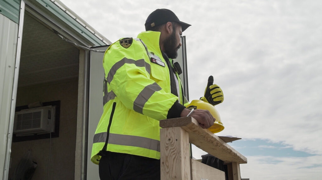 A person in a high-visibility jacket stands by a wooden railing, holding a yellow hard hat and giving a thumbs up near an open door.