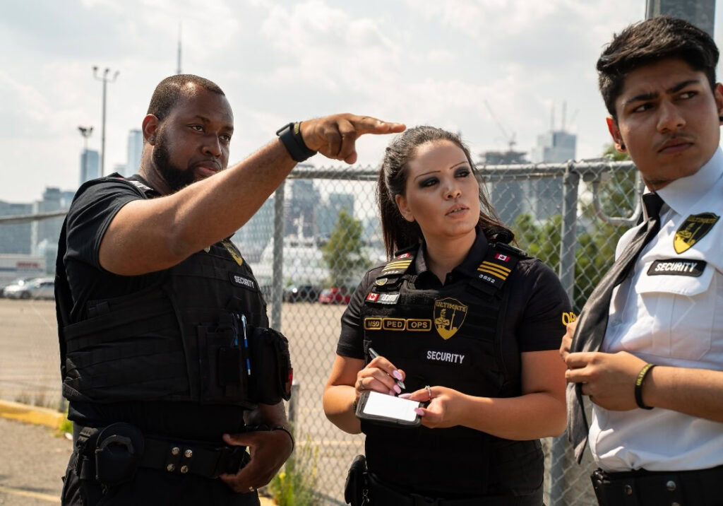 Three security guards stand outside by a chain-link fence; one points in the distance while another, representing security management services, writes on a notepad, and the third looks away.