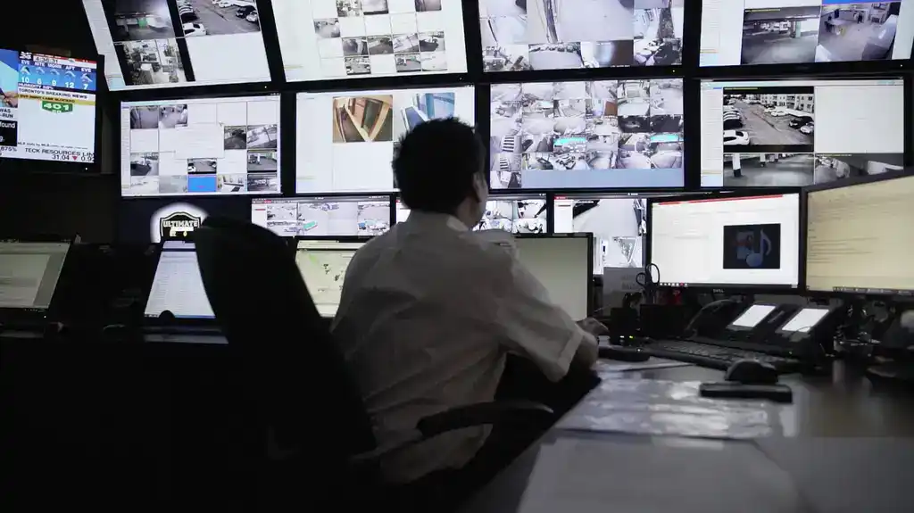 A person in uniform monitors multiple surveillance cameras and computer screens in a control room, overseeing live security footage and data displays as part of professional security services in Toronto.