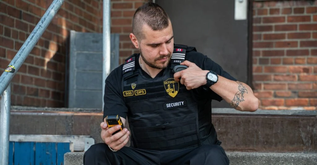 A security guard in uniform, responsible for parking enforcement, sits on outdoor steps holding a radio and checking his watch, with a brick wall behind him.