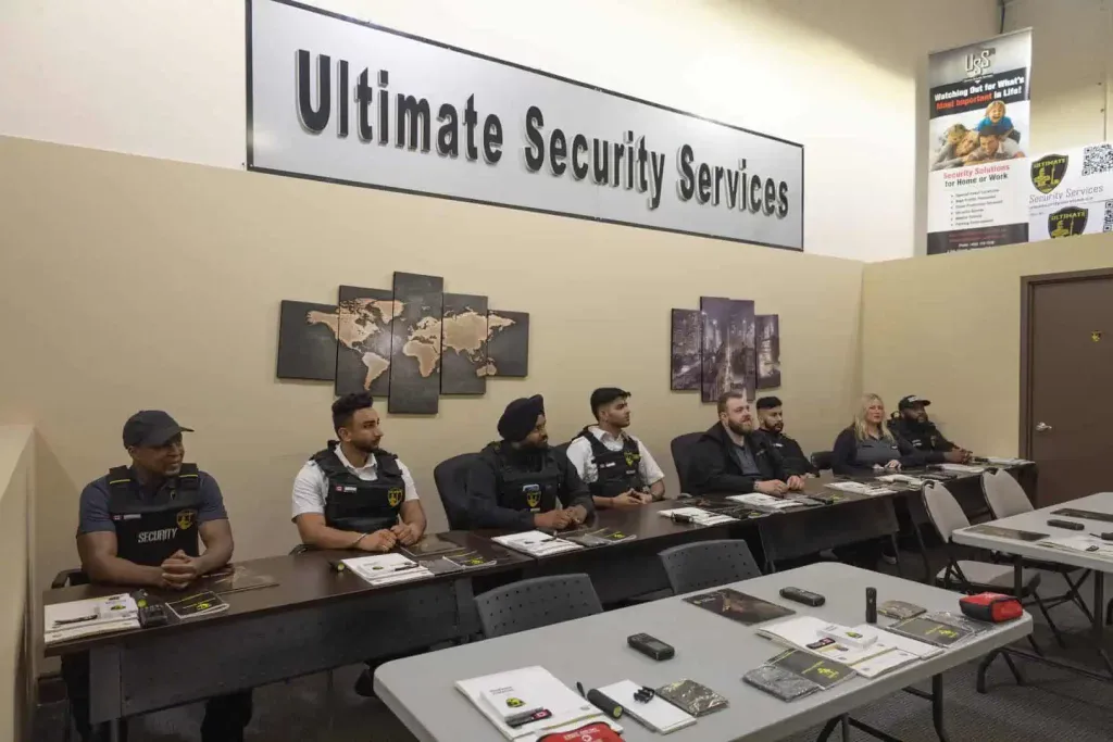 A group of security personnel in uniform sit at desks in a training classroom under a sign that reads "Ultimate Security Services," one of the top security companies in Toronto.