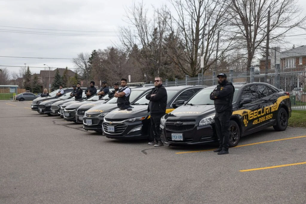 Seven security personnel stand beside a row of black security cars parked in a lot on an overcast day, highlighting professional security guard services in Toronto, with bare trees and a metal fence in the background.
