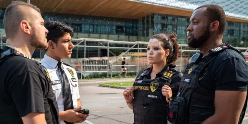 Four security guard company personnel stand in a group outdoors, engaged in conversation, with a modern building and scaffolding in the background.