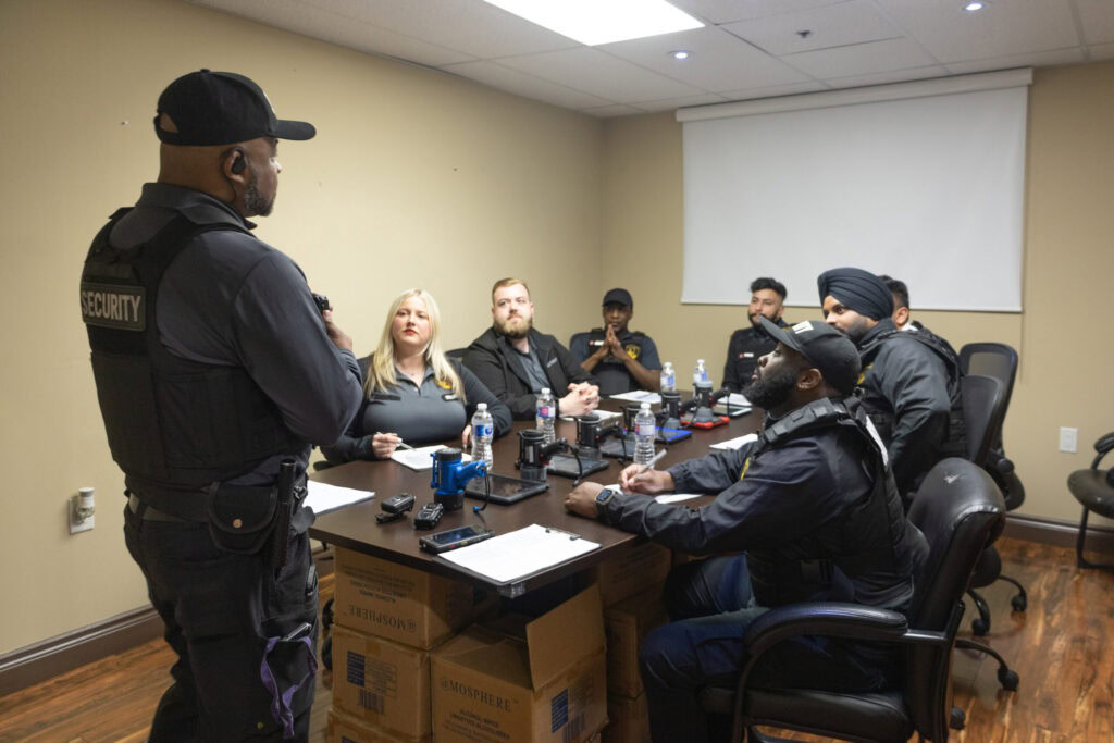 A security officer stands and speaks to six seated colleagues around a conference table in a meeting room equipped with notepads, radios, and water bottles—exemplifying the professionalism found at top security companies in Toronto.
