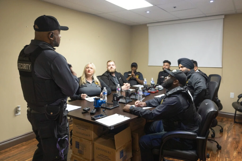 A security officer stands and speaks to six seated colleagues around a conference table in a meeting room equipped with notepads, radios, and water bottles—exemplifying the professionalism found at top security companies in Toronto.